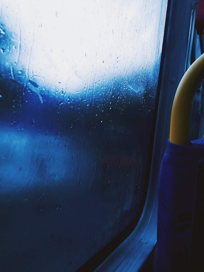 Raindrops on a bus window create a moody, abstract scene with blurred blue hues.